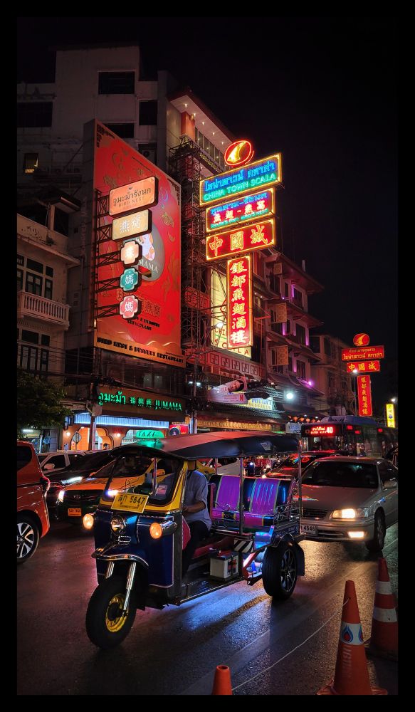 Nighttime street photography in Bangkok, Thailand. A tuktuk in the street surrounded by cars, with neon signs in the background.
