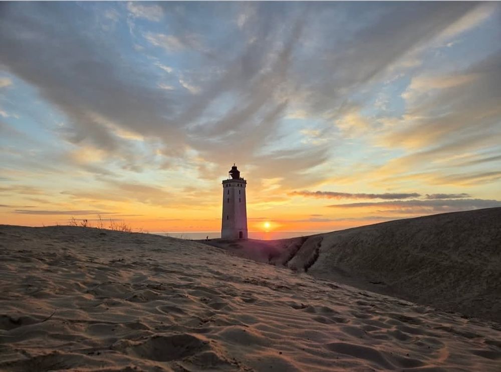 Die untergehende Sonne in den Dünen - ein Leuchtturm in der Bildmitte. 