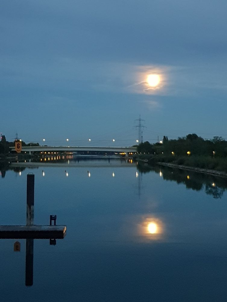 ​Es zeigt eine Nacht- oder Dämmerungsaufnahme eines Sees unter einem dunkelblauen Himmel. Ein heller Vollmond spiegelt sich auf der ruhigen Wasseroberfläche. Im Hintergrund ist eine Brücke mit Beleuchtung sowie ein Strommast und Bäume zu sehen. Im Vordergrund ragt ein kleiner Steg mit einer Bootsanlegestelle ins Wasser.
