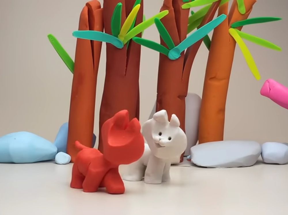 Screenshot from childrens clay animated series Mio Mao, showing one red and one white cat made of clay, standing on all fours, the background made up of very simple clay blobs representing stone and trees