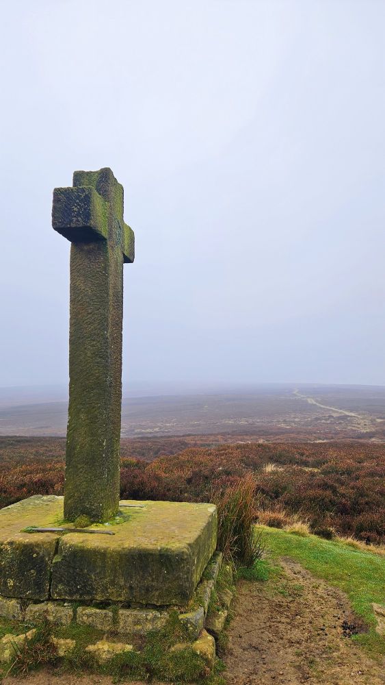 To the left in the foreground is a stone cross waymarker from the early 1800s, the stone is green due to a light layer of moss. Behind the cross, is moorland covered in brown heather spreading far into the distance. The sky is white with thick mist.