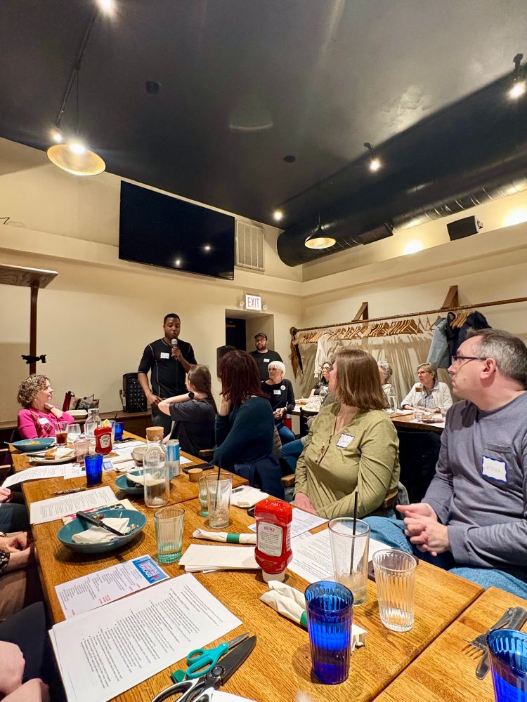 Group of people at 2 long tables listening to a speaker.