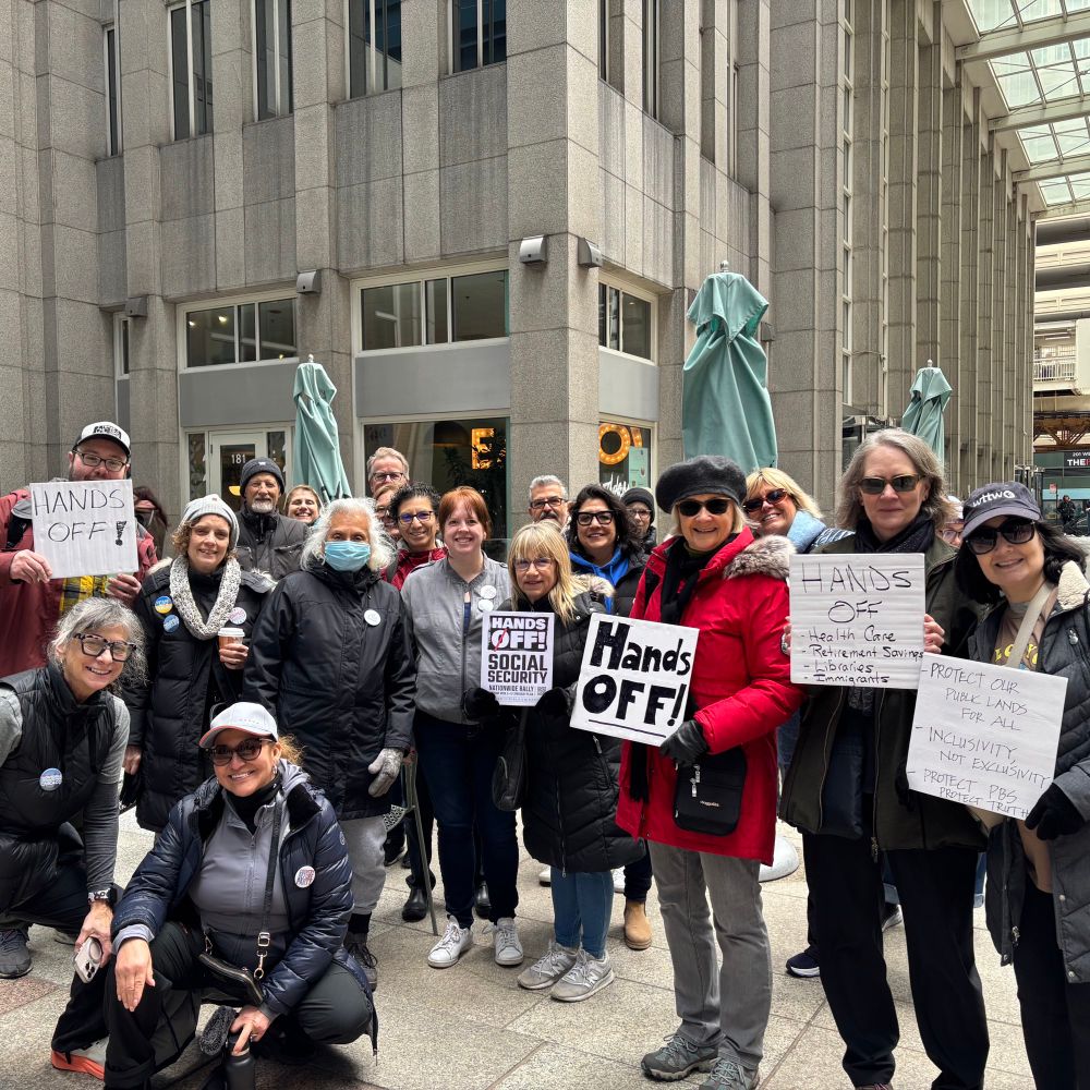 Group of nearly 20 people standing in front of a coffee shop holding various protest signs, largely with a “Hands off!” message