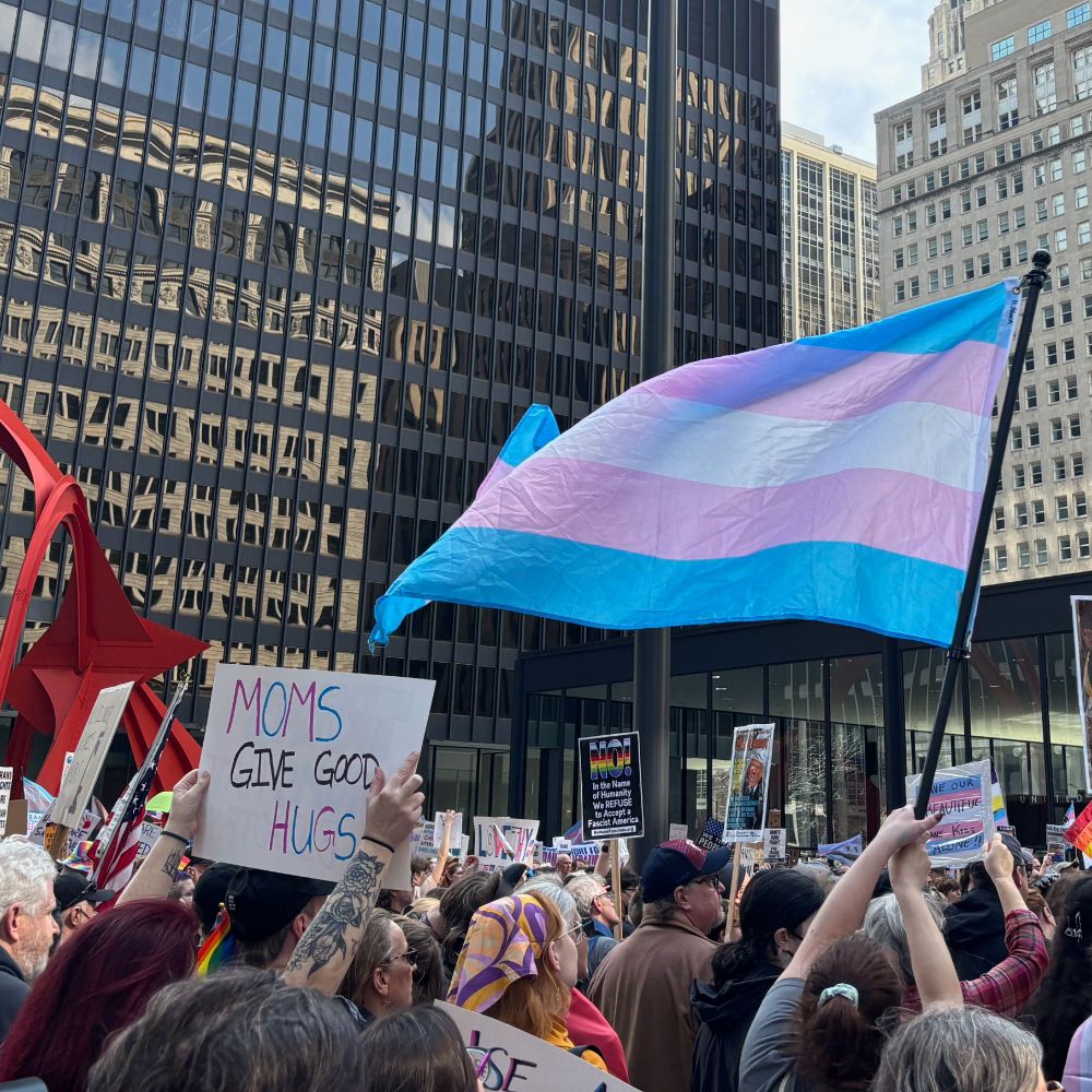 Trans flag flying above the crowd. Many signs can be seen, including "Moms give good hugs" in trans pride colors