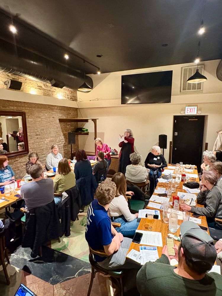Group of people at 2 long tables listening to a speaker.
