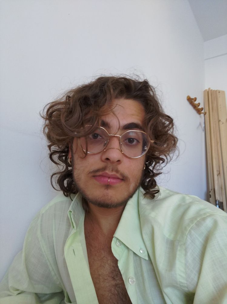 Moi, un jeune homme de 20ans, au cheveux brun bouclé mi-long et aux yeux brun. J'ai la peau bronzée et une petite moustache et barbiche. J'ai un labret, une nose chain, des lunettes rondes et plusieurs percings aux oreilles. Tout ces bijoux sont de couleur dorée. Je porte une chemise ouverte, de couleur verte pâle tirant un peu sur le jaune.