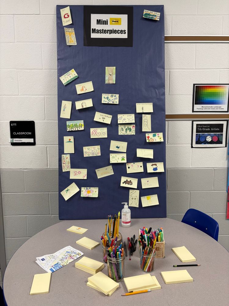 Interactive station called Mini Masterpieces encouraged visitors to make small art. Table in the foreground has sticky notes and art supplies. In the background is a piece of black butcher paper with sticky note art displayed. 