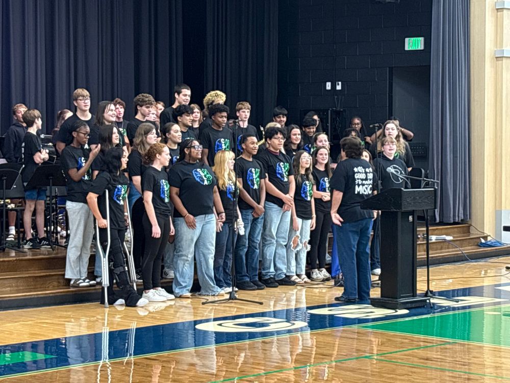 A small choir of high school students performs in a school gym. They are dressed in jeans and black tshirts. The director stands in front of them, back the camera. The back of her shirt says, “It’s a good day to make music!”