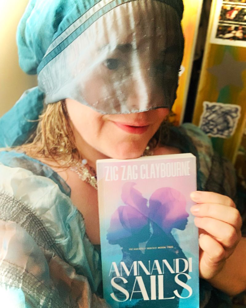 Woman in teal gauze veil and dress holds up a paperback book of similar colors. The book is AMNANDI SAILS by Zig Zag Claybourne.