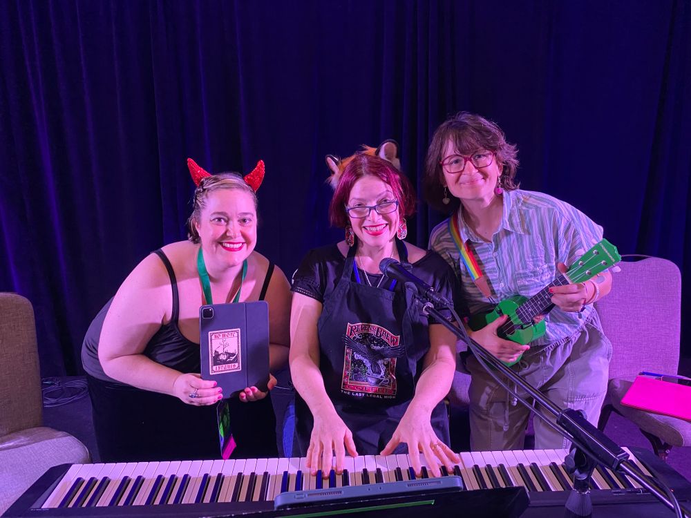 Left to right: C. S. E. Cooney, holding an iPad with a sticker that reads "No More Tyrants" and wearing a pair of devil ears. Dr. Mary C. Crowell, wearing an apron and a pair of cat ears, playing a keyboard. Tina Connolly, playing a green ukulele on a rainbow strap. The picture was taken after a concert they did at Convergence 2025, in Minneapolis, Minnesota: the first time they played music from "The Devil and Lady Midnight" for the public.