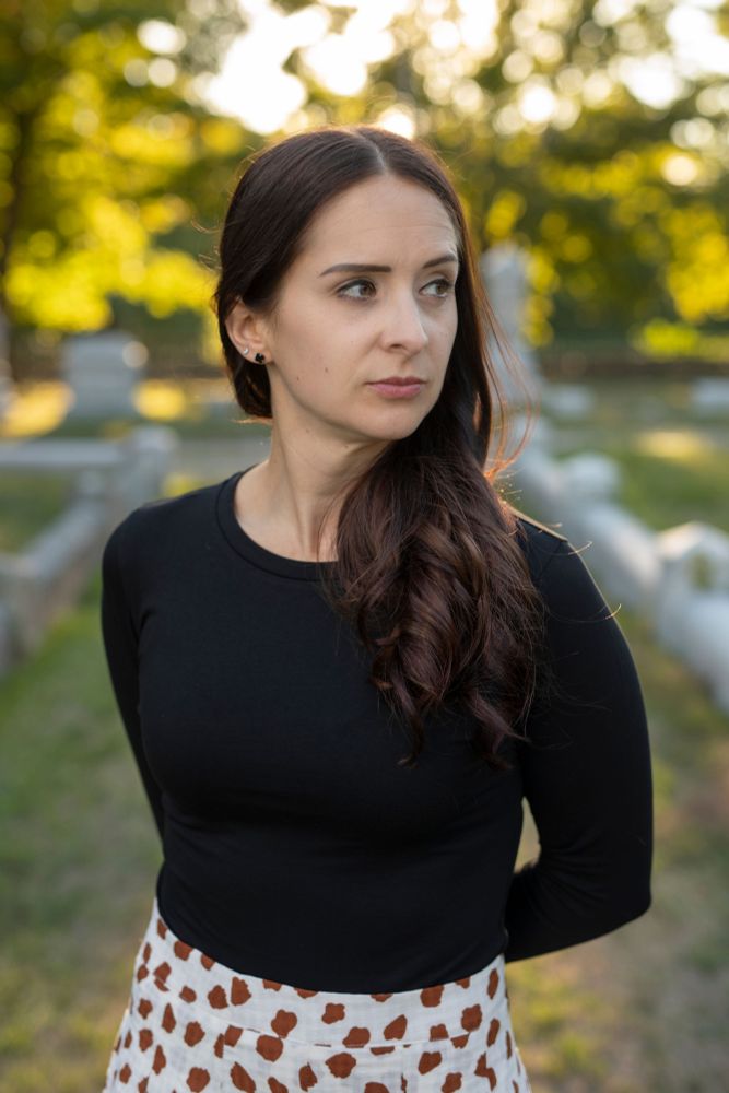 Author pic of Christa Carment: an Italian-American woman with brown hair and brown eyes, wearing a black shirt, and white-and-red patterned skirt, looking off into the distance against a backdrop of a green graveyard.