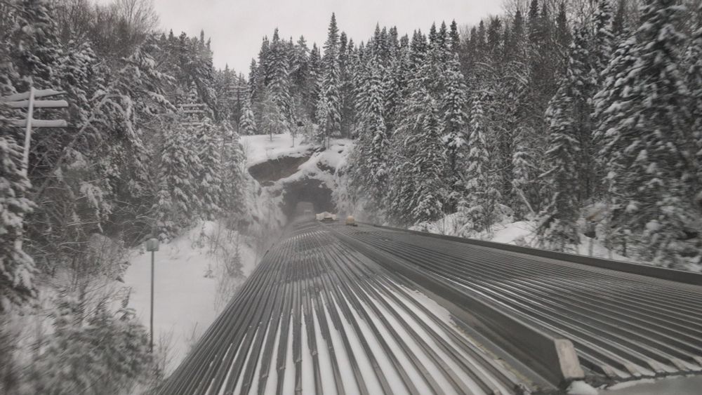 Rear-facing view from the Dome Car as the VIA Rail Canadian train exits a tunnel on the CN Redditt Subdivision in a remote area of northern Ontario. The tunnel cuts through a rocky ridge surrounded by dense, snow-covered coniferous trees.