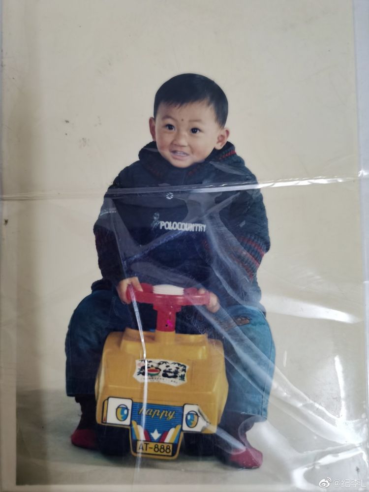 baby photo of ji li. hes dressed in a very warm woolen sweater and sitting astride a plastic toy car. he's got a kind of intense expressiom on his face and looks like he's already a whole handful. 