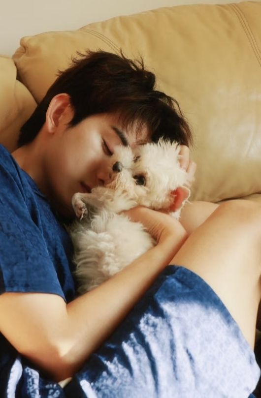 photo of actor zhu zanjin cuddling his white poodle wooly on a beige sofa. he is wearing some sort of short-sleeve & short pyjama set and is curled up into a little ball around the dog. plenty of thigh on view.