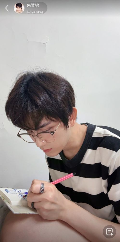 Screenshot of Zhu Zanjin's live. He is bent over a tiny notebook that he is holding on his bare knees and focused on drawing a flower. He is wearing wire-rimmed glasses, a tshirt with broad, black-and-white stripes and shorts so tiny they are invisible in the picture. He has rings on the index finger and the ring fingers of his drawing hand.