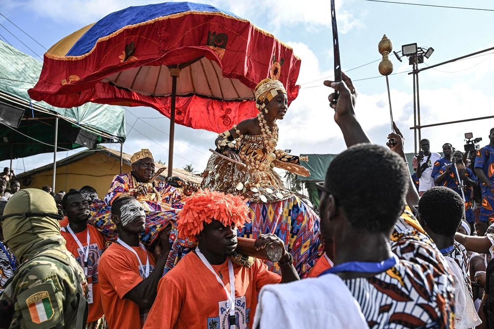 Nanan Awoulae Desire Amon Tanoe, king of the N'Zima people of Grand Bassam and president of the chamber of kings and traditional chiefs of Cote d'Ivoire (CR) carried by porters in a pirogue with a girls dancing in traditional clothes, greets supporters as he arrives for the celebration of Abissa festival in Grand-Bassam on October 18, 2025. Abissa is an annual festival celebrated by the NíZima Kotoko people in Grand-Bassam, a UNESCO World Heritage site. (Photo by Sia KAMBOU / AFP)