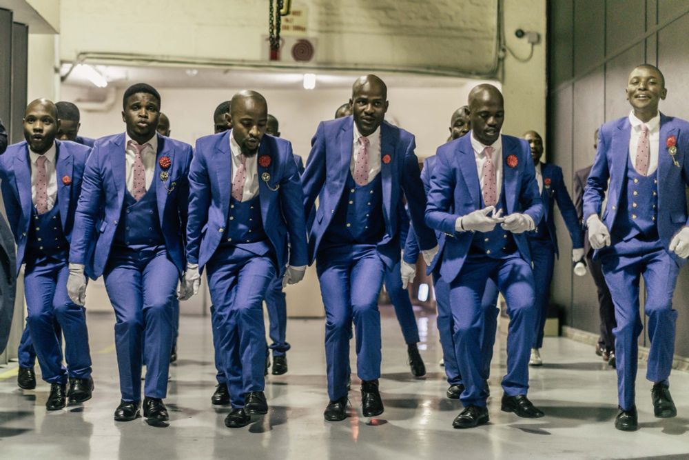 A group of sharply dressed men in matching royal blue suits stride in unison down a corridor, their movements precise and graceful. They wear pink polka-dot ties, red lapel flowers, and white gloves, exuding both confidence and elegance as they prepare to perform. 