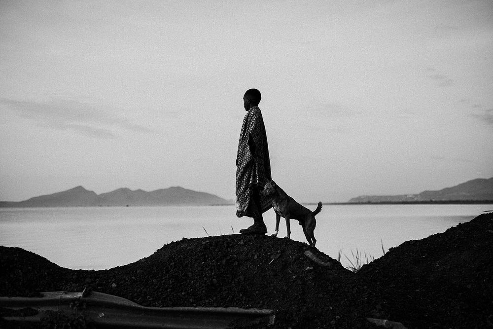 The 14th edition of the Uganda Press Photo Award (UPPA), the 8th edition of East African Photography Award (EAPA) have just completed. Lucas Oyugi was one of the photographers recognised for this photograph.
 At the shores of Lake Abaya in southern Ethiopia, a young boy and his dog stand silently—wrapped in warmth, surrounded by stillness. In this quiet moment, their presence becomes one with the land and water. This image speaks to a deep, unspoken bond between human, animal, and environment—where companionship becomes guardianship, and solitude feels sacred. It's a story of belonging, of being rooted without words.