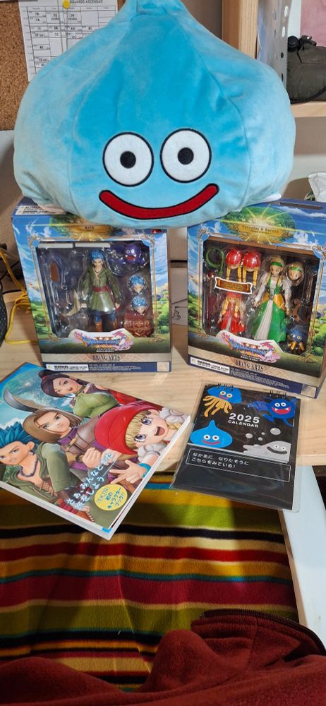 Picture of all the merchandise received. A Slime plushie, Bring Arts figurines of Erik, Serena and Veronica, DQ11 character book and the official monster DQ calendar of 2025.