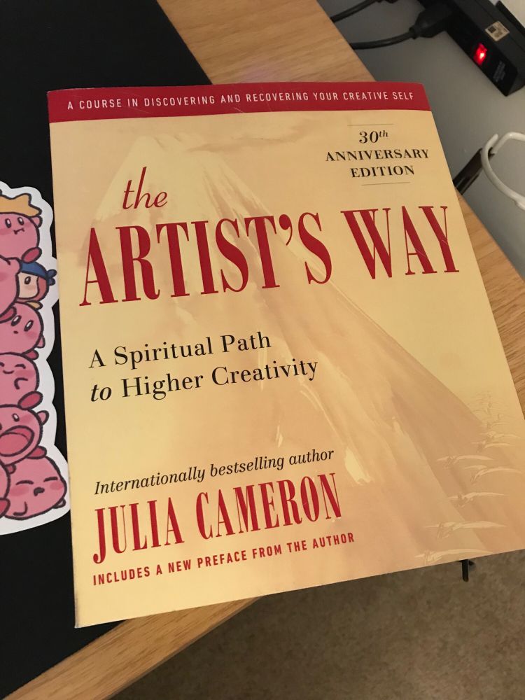 A copy of The Artist’s Way book by Julia Cameron on a wood desk, next to a kirby bookmark.