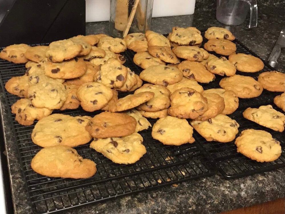 Roughly 60 chocolate chip cookies on black drying racks on top on a gray granite counter