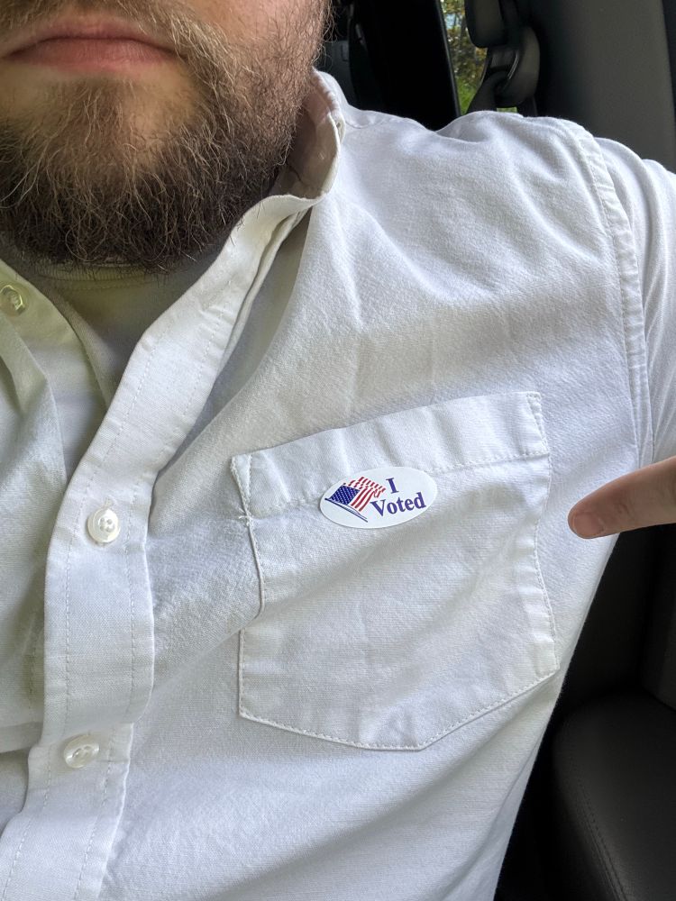 Man wearing white shirt pointing to an “I Voted” sticker