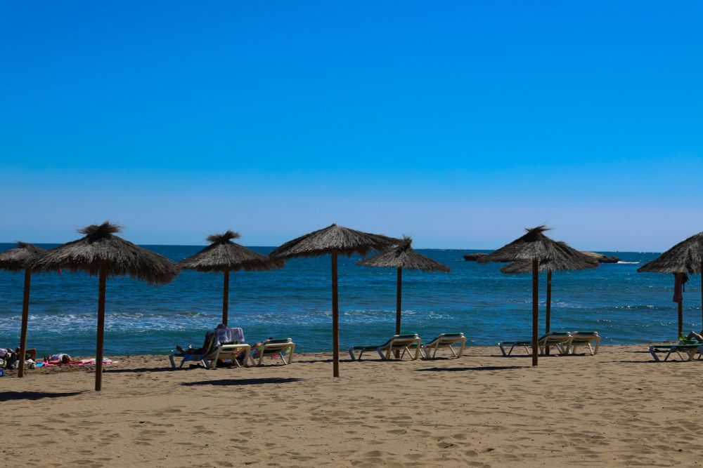 Zu sehen sind verschiedene Strohschirme am Strand, im Hintergrund das Mittelmeer.