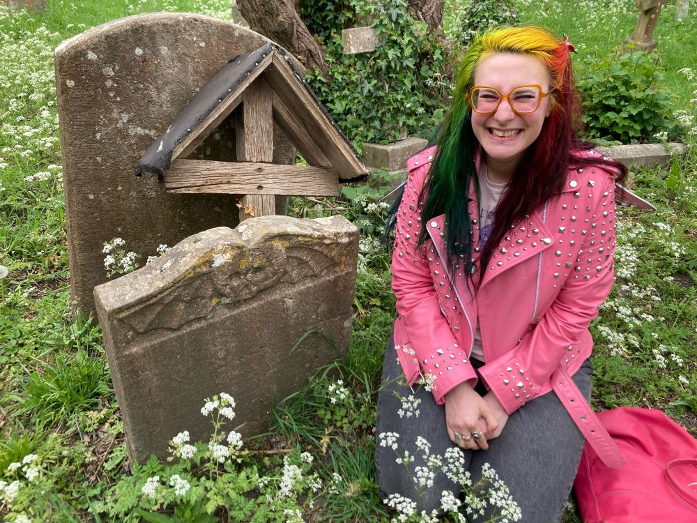 A woman with rainbow hair and a pink biker jacket sitting next to a headstone with a cranium flanked by bat wings on the upper portion