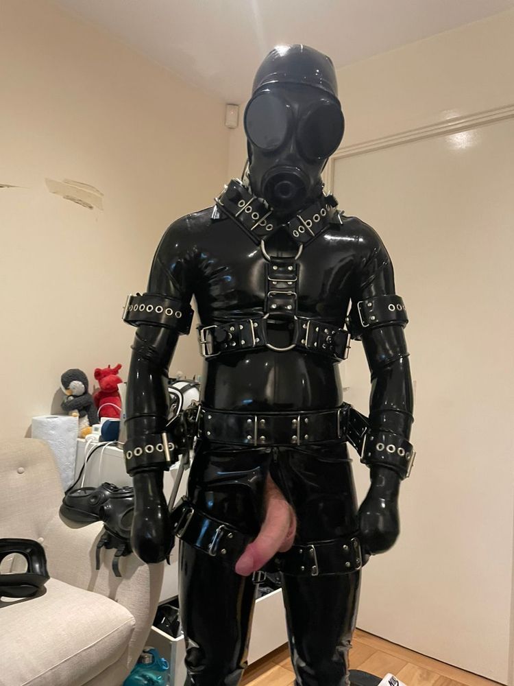 A drone in a catsuit, body harness, cuffs, gasmask and mitts, all in black rubber, with his semi-erect dick hanging out