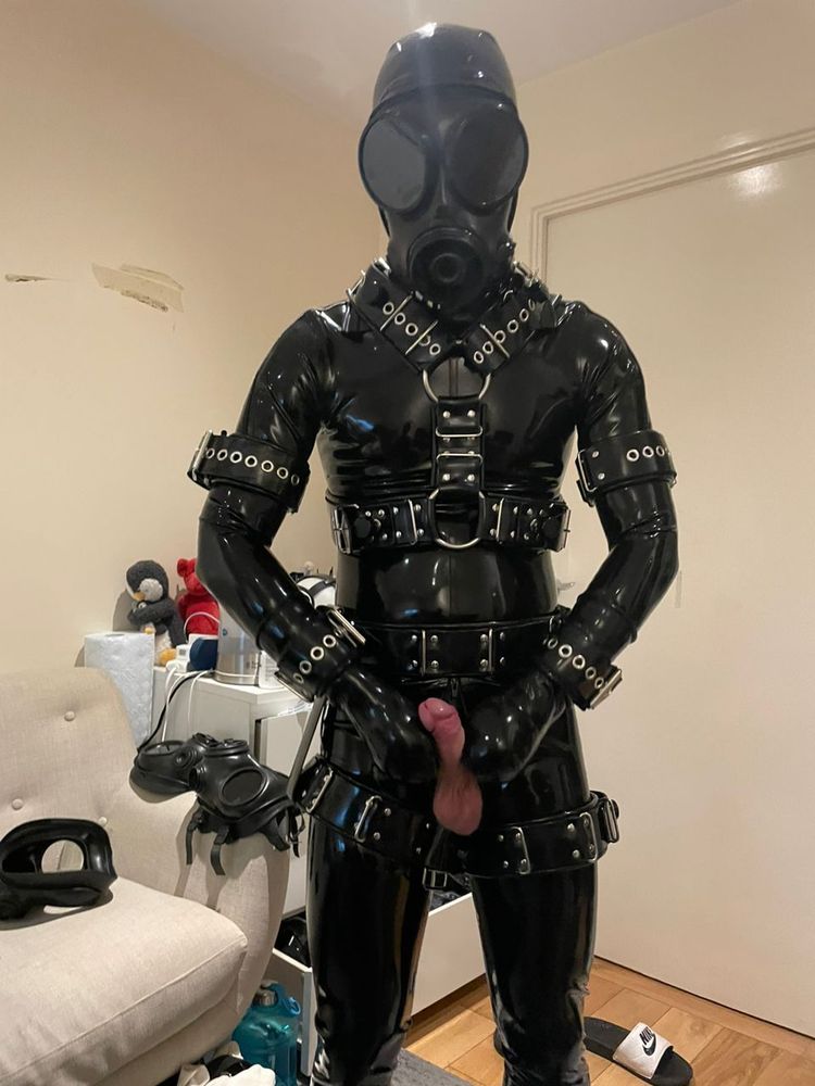 A drone in a catsuit, body harness, cuffs, gasmask and mitts, all in black rubber, holds his erect dick
