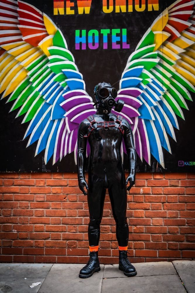 Drone in black latex catsuit, gas mask, boots and harness in front of an angel wing mural in Manchester