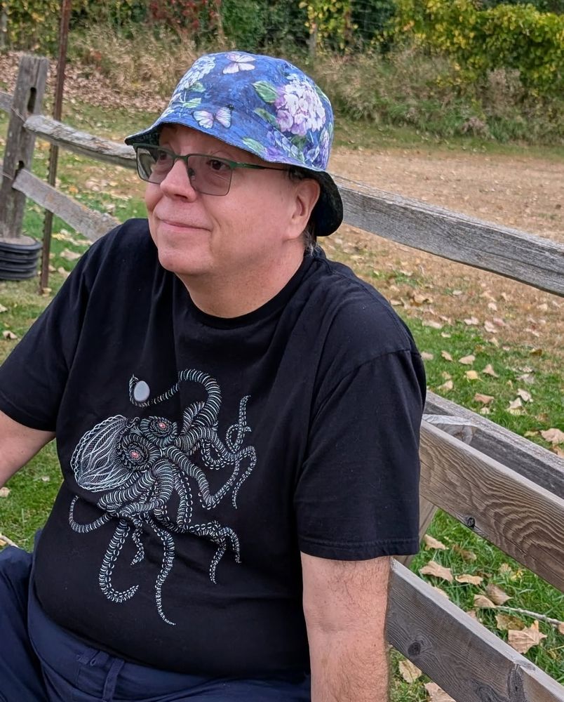 photo of white guy with tinted glasses, blue floral bucket hat and wearing a black t shirt with red- eyed squid holding a pearl. He is sitting against a wood fence leaning on left arm