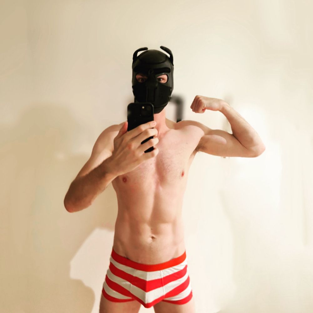 A shirtless male model wearing a black puppy hood poses in front of a mirror, flexing his arm confidently. He is wearing festive red-and-white striped briefs, evoking a candy cane vibe, against a simple background. The playful and confident tone matches the holiday spirit.