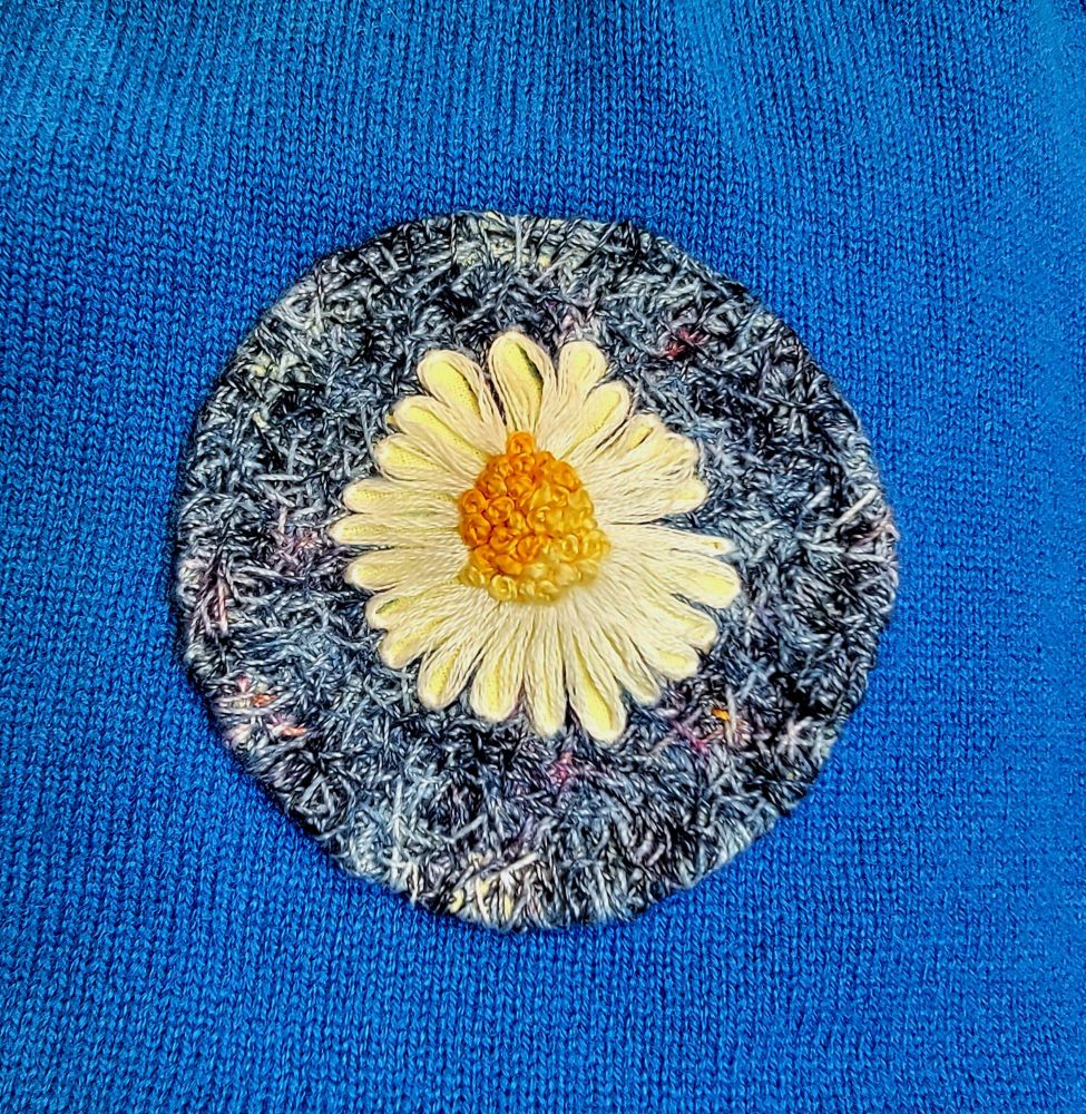 Photo of a blue cashmere sweater with a patch attached. The patch is a hand embroidered daisy with a randomly stitched background in hand dyed indigo threads.