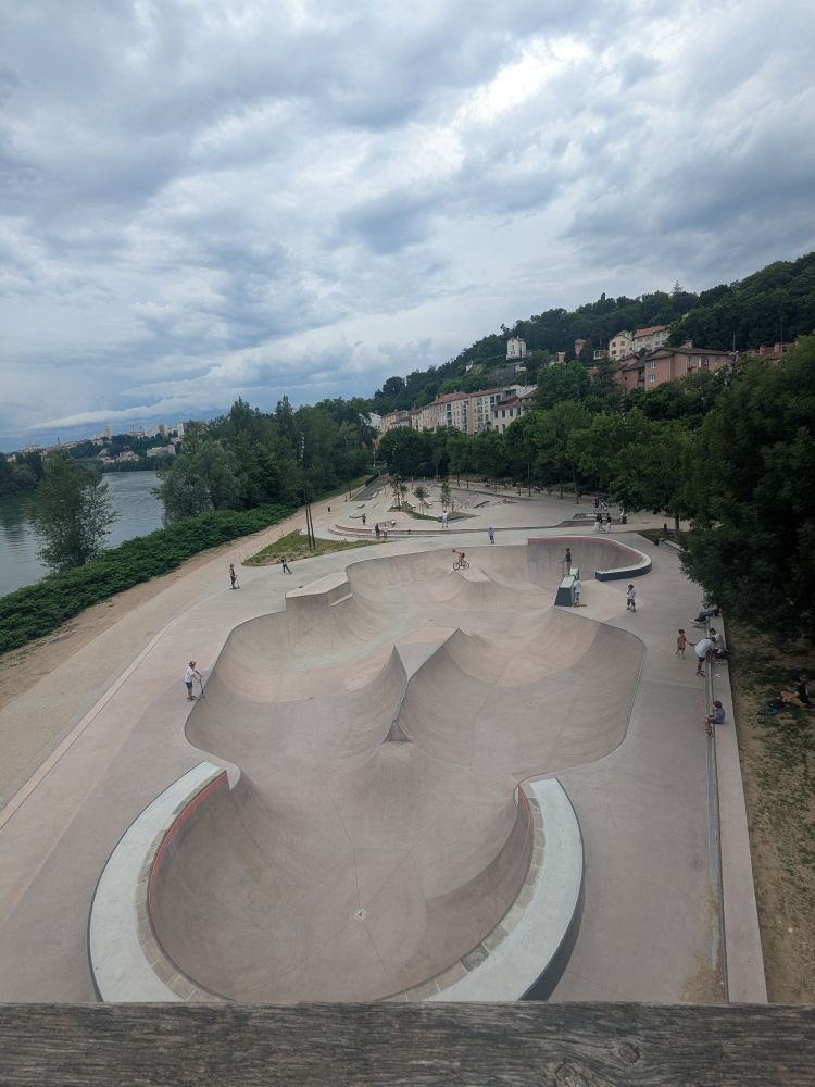 Vue sur un skatepark tout neuf (creusé en béton) au bord du Rhône.
Bordé d'arbres et du Rhône. En arrière plan : la colline de la Croix-Rousse à Lyon. 