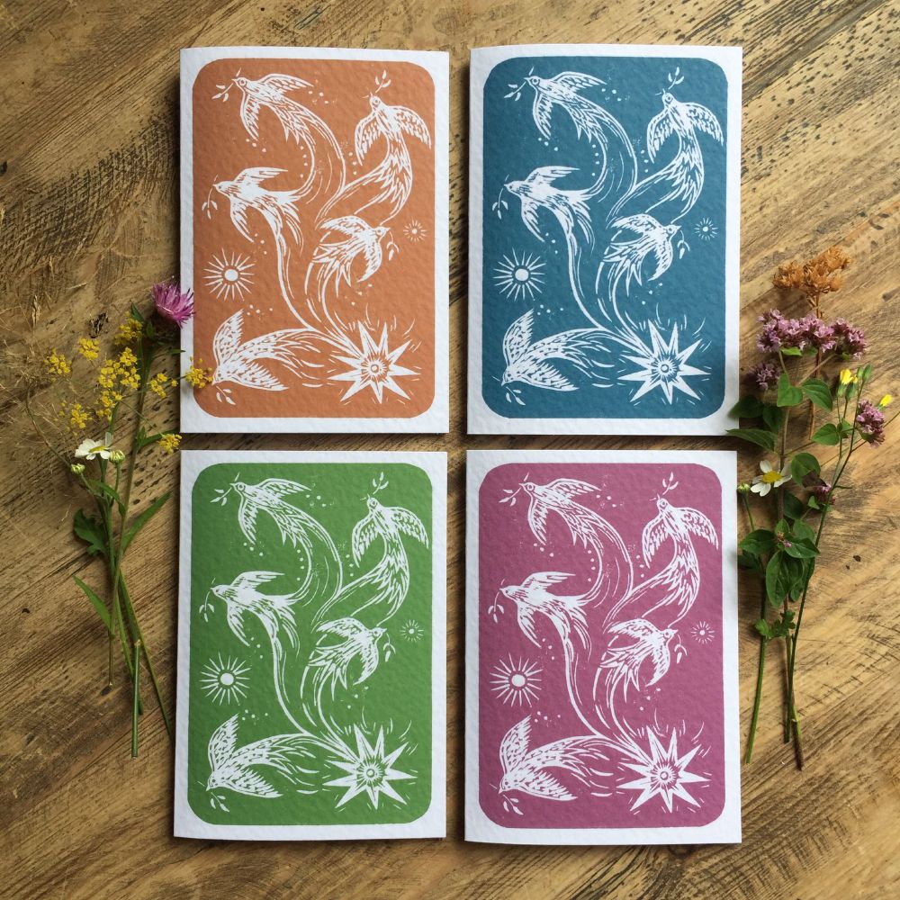 This is a photograph of some greeting cards on a wooden table - they have flowers on either side of the group and they are in 4 different colours : Rust, Teal, Green and Plum. They are reproduced digitally from my original linocut which I hand carved a few days ago. The subject is of a shooting star from which birds of peace appear to be swirling out into the sky.