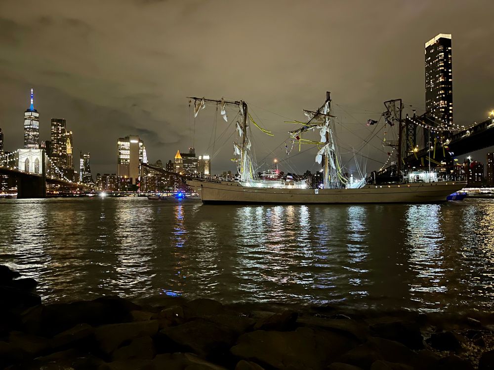 A tall ship between the Manhattan and Brooklyn Bridges at night with its topmasts broken