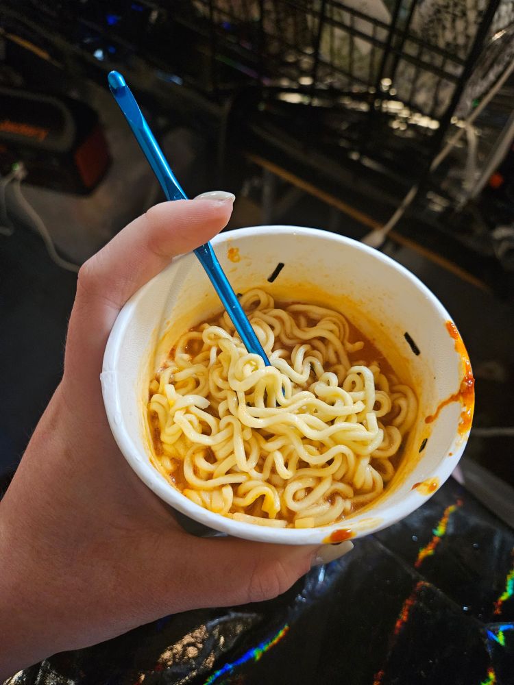 Photo of a hand holding a cup noodle with a blue crochet hook sticking out of the noodles. The sauce is red-orange.