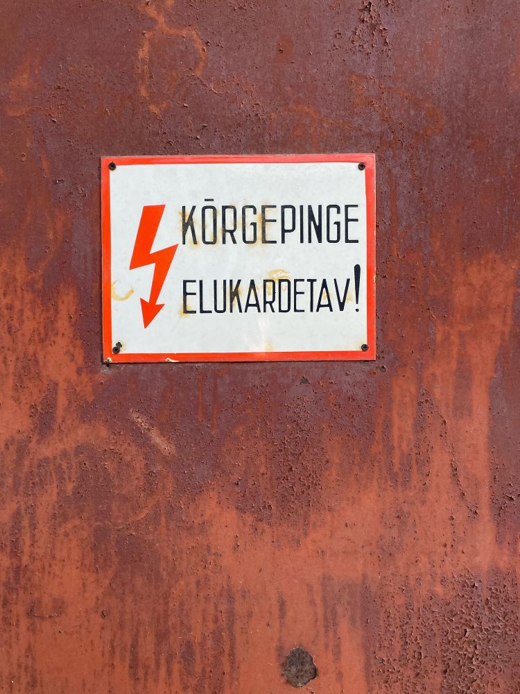 A vintage health and safety sign, in tall lettering and written in Estonian, saying ‘Korgepinge Elukardetav!’ With a lightning bolt symbol. The red lettering and lightning suggest danger. 