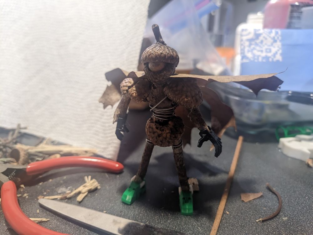 A character made of mostly acorns and wires stands on a table, the character also has a cape made of a dried leaf. There is a paper napkin behind him and a lot of broken pieces of wood and a Miller and scissors in the background.