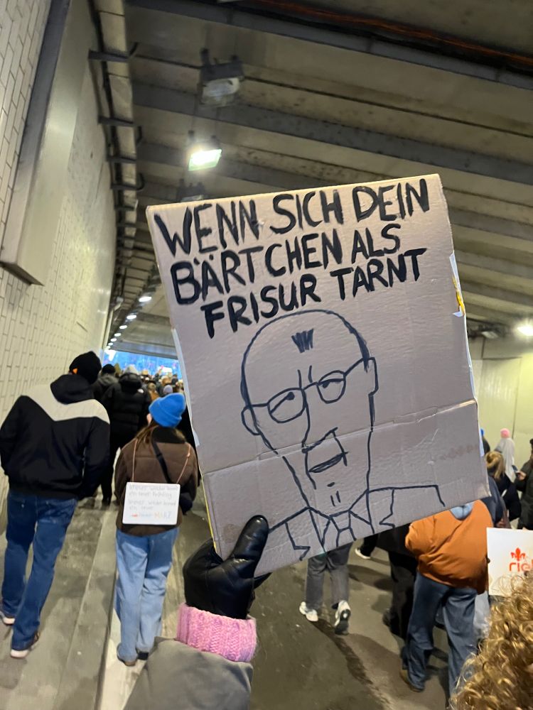 Selbstgemachtes Plakat zeigt Merz mit seinem charakteristischen Haartest auf der Stirn und dem Text: Wenn sich das Bärtchen als Frisur tarnt.