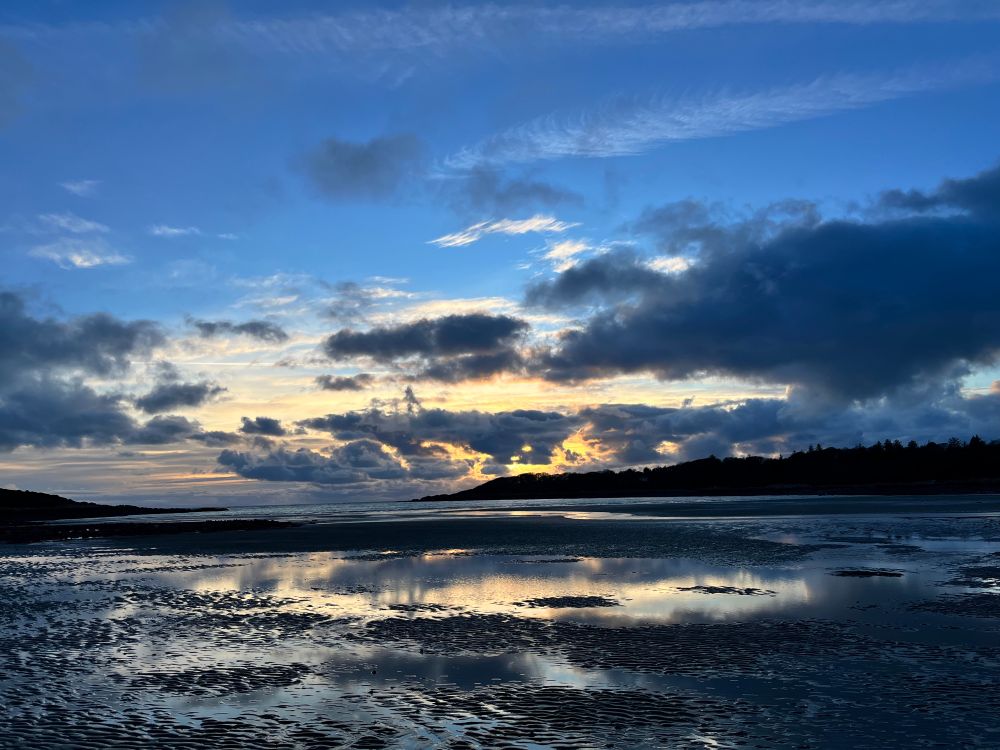 Light reflecting on patches of water on the beach. Blue sky above grey clouds.