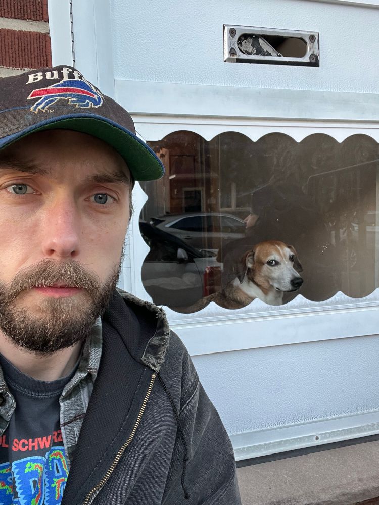 A selfie of me sitting on the stoop in the foreground while Milo (beagle dachshund mix) stares out, looking annoyed, through the storm door