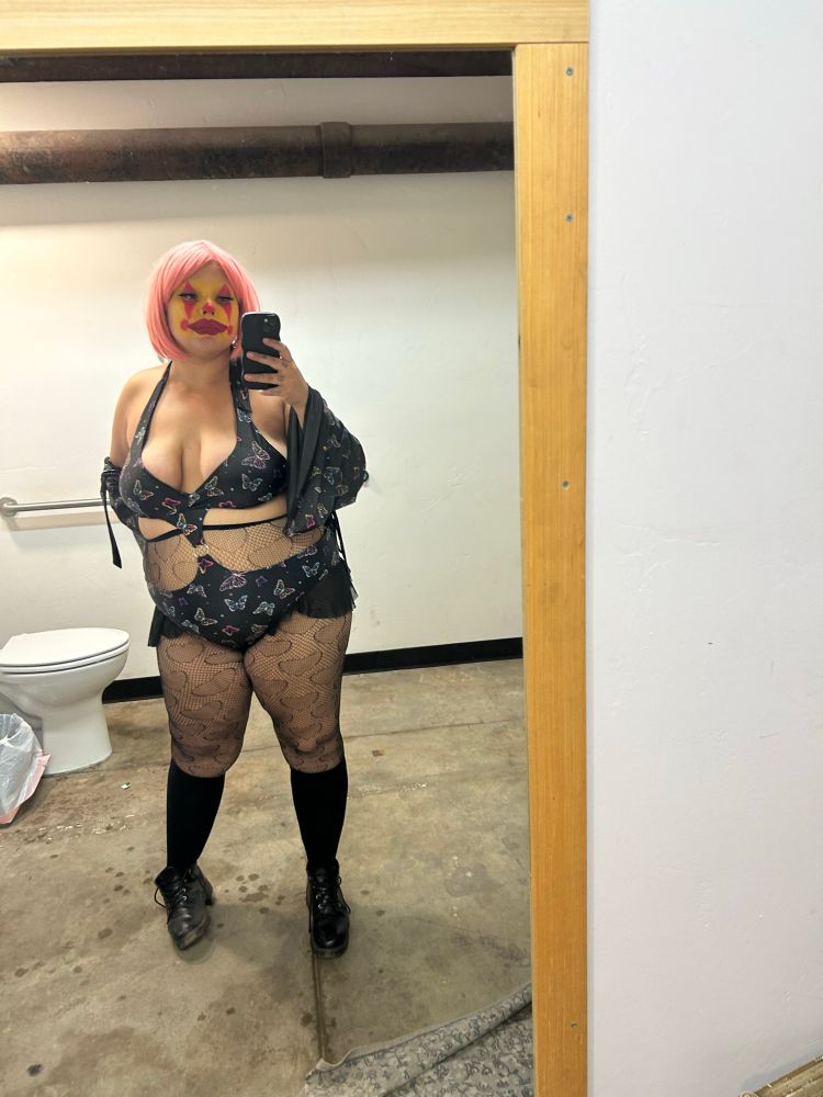 Anya stands in front of an almost floor to ceiling bathroom mirror in a light pink wig and yellow and hot pink clown makeup wearing a butterfly print halter top bikini-esque rave outfit connected in the mid section by a metal ring. The bottom half has mesh flaps that imitate a mini mini miniskirt. They are also wearing matching flowy sleevlettes, heart-patterned fishnets, black knee high compression socks, and untied dusty combat boots.