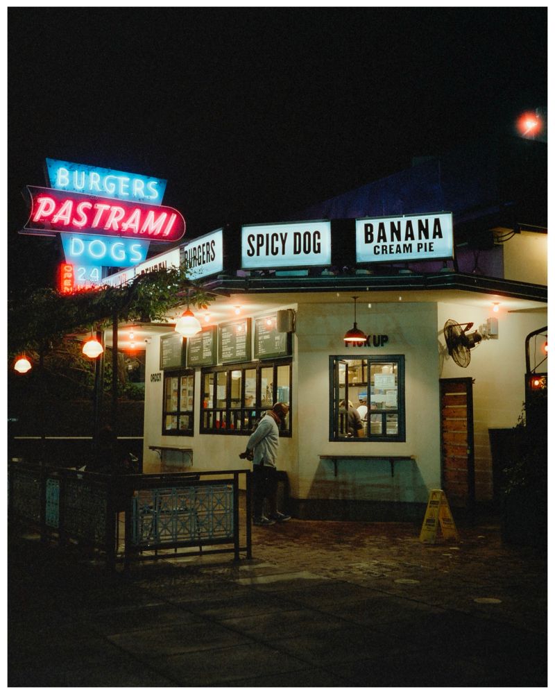 This image shows a cozy and inviting late-night food stand or diner with a retro vibe. The signage highlights menu items such as burgers, pastrami, hot dogs, and a “spicy dog,” along with a dessert option, “banana cream pie.” Neon and illuminated signs create a nostalgic, warm atmosphere. The stand has a pickup window with a customer waiting, and small, glowing red lamps add charm to the outdoor seating area. The night setting enhances the scene’s relaxed and urban feel.