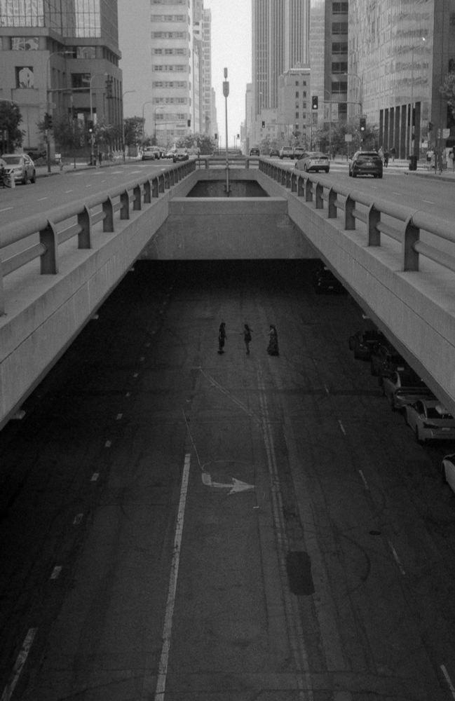The image depicts a black-and-white urban scene featuring a sunken roadway beneath a street-level bridge in a cityscape. The perspective is taken from a higher vantage point, looking down at the underground road, which has visible lane markings, including an arrow pointing forward.

Above the tunnel, the city streets are active with cars, buildings, and pedestrians walking along the sidewalks. The architecture consists of a mix of modern and older high-rise buildings, suggesting a dense metropolitan area.

In the lower part of the image, three individuals are seen standing in the underground roadway, their figures appearing small and slightly obscured by darkness. Their presence creates an eerie and mysterious atmosphere, as they appear to be engaging in some interaction in an otherwise empty, desolate space.

The black-and-white composition enhances the stark contrast between the brightness of the upper street level and the darkness of the lower road. The grainy texture and lighting contribute to a cinematic and somewhat surreal quality, evoking a noir or dystopian feel.