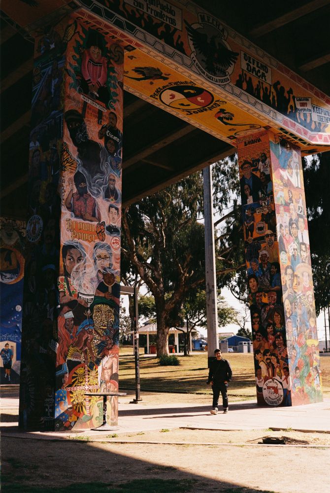 This image captures a vibrant outdoor mural painted on large concrete pillars, likely under a bridge or overpass. The murals depict intricate and colorful artwork reflecting cultural, historical, and social themes. They include portraits, symbols, and text celebrating identity, community, and activism, with phrases like “Brown Power” and imagery of influential figures and symbolic designs.

The setting appears to be a park or community space, with trees, a playground, and a person in dark clothing standing nearby, observing the artwork. The light and shadow interplay enhances the visual impact of the scene, emphasizing the richness of the murals. This appears to be a space celebrating cultural heritage and resilience.