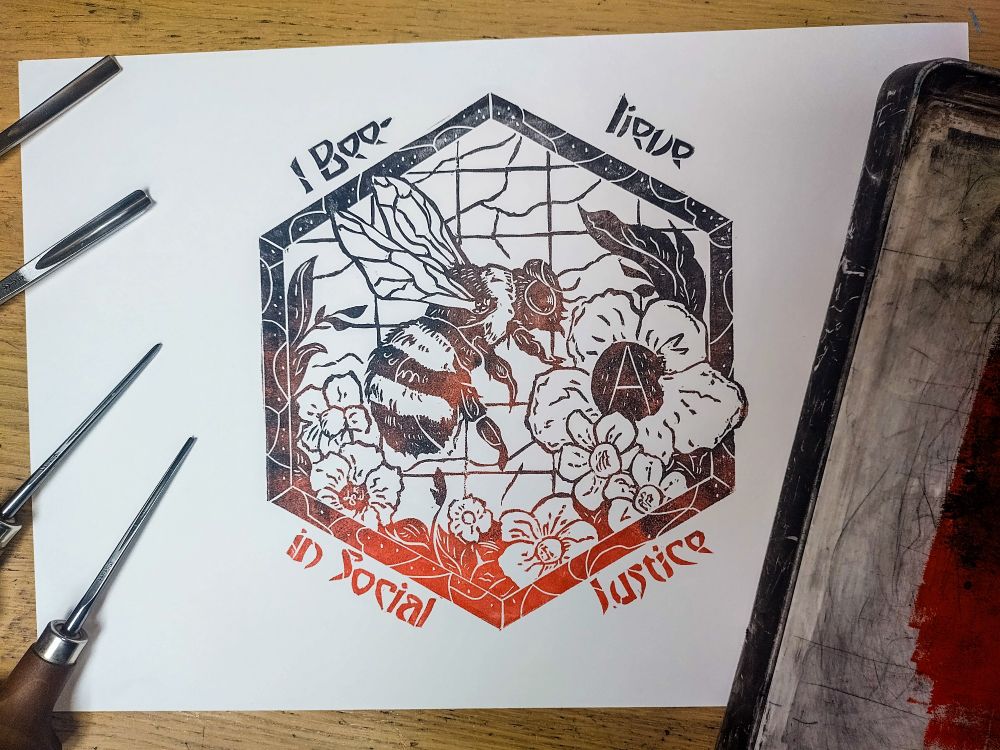 Une impression linogravure en dégradé de noir et rouge et dans un style Art Nouveau. Un gros bourdon à casquette vole au milieux des plantes et s'apprête à butiner une fleur symbolisant l'anarchie.
L'impression est placée entre des gouges et un plateau encreur. 