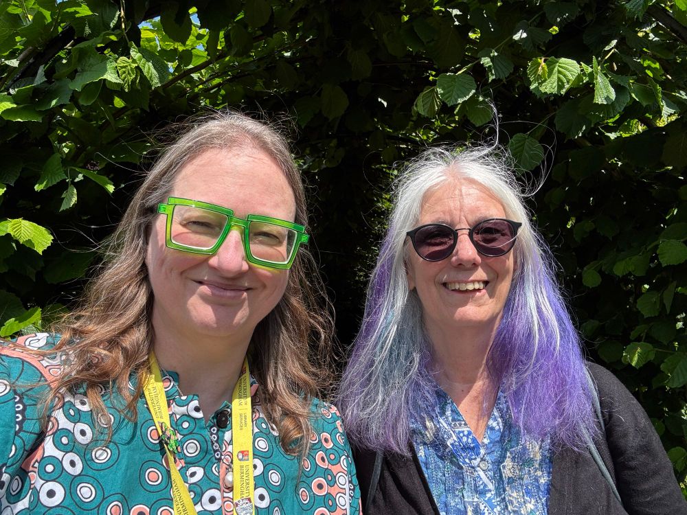Danielle (left) and Mary (right). Mary has epic long hair that is white, purple and blue. She is wearing a blue top. Danielle has comic effect green glasses and a Zuri African dress that has a pattern that looks like Covid. 