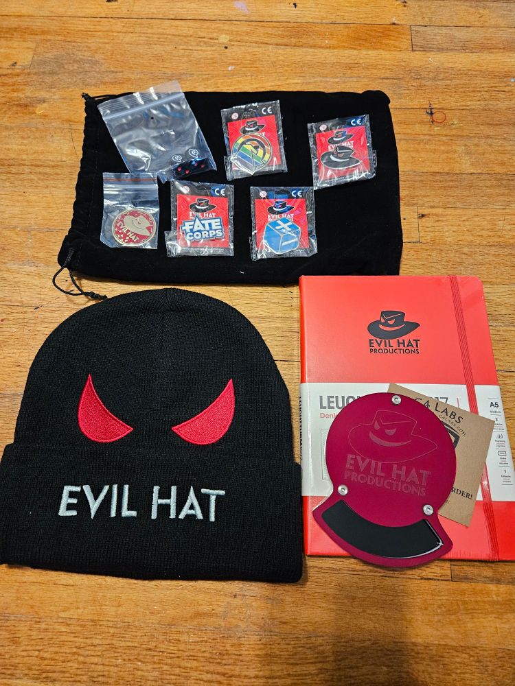 A collection of support from Evil Hat Productions including:
A large black bag
A black beanie with red eyes that says "Evil Hat"
A red notebook that has the Evil Hat logo on the front.
A red coaster from C4 Labs with the Evil Hat logo
A paid of d6 with the Evil Hat as the 6
A metal d2 with a red side that says "Evil Hat" and has the Evil Hat on it (the reverse is blue says "Good Hat" and has the Hat smiling.
The Evil Hat pin
A blue Fate d6 pin with a blank side, plus side, and minus side showing
A Fate Corps pin
A pin with a rainbow circle and a shape that represents a d6 rolling by using an arrow looping over it
